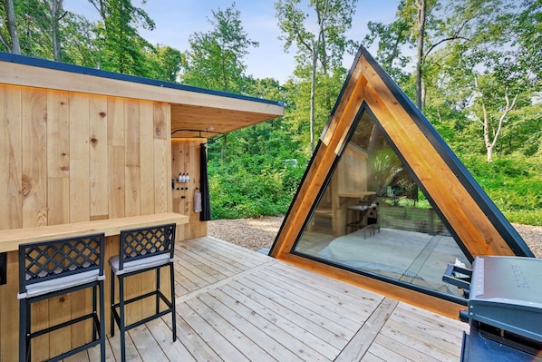 Une cabane en bois avec une extension triangulaire en verre, une terrasse avec deux chaises et un barbecue.