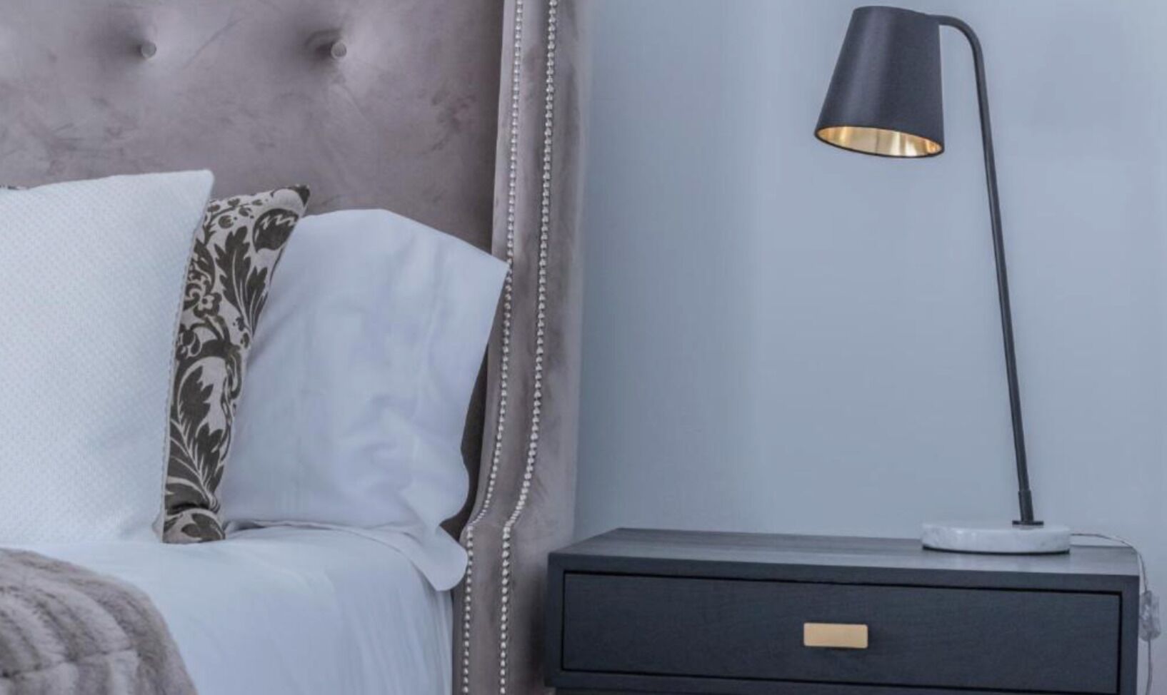 A bed with white and patterned pillows, a grey headboard, a black bedside lamp, and a dark nightstand with a drawer.