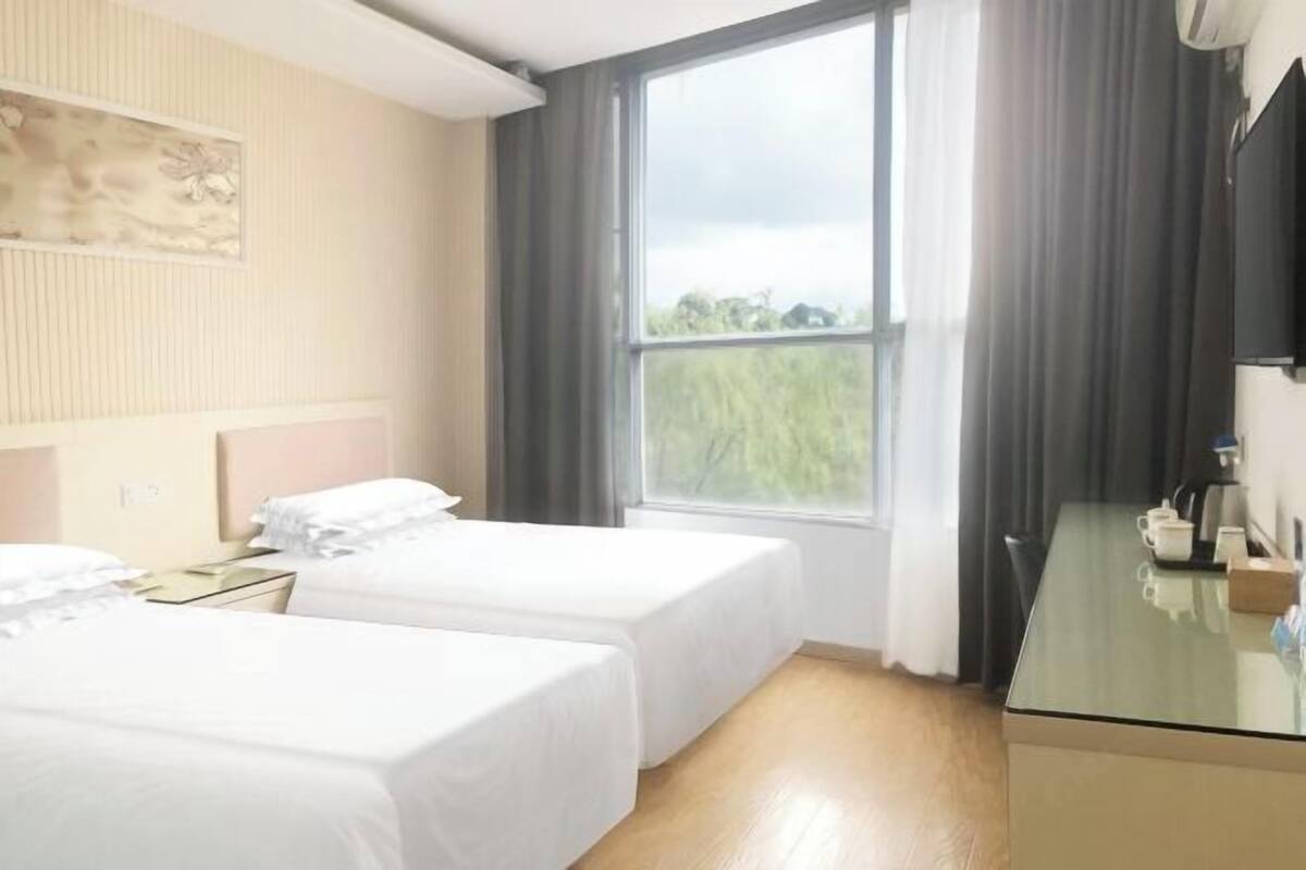Ein Hotelzimmer mit zwei Einzelbetten, einem Glastisch, einem Flachbildfernseher und einem großen Fenster mit Vorhängen.
