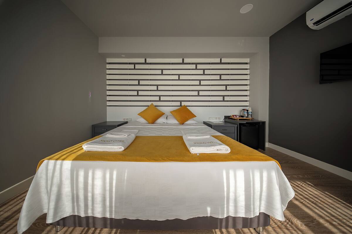 Cama bem arrumada com colcha branca e duas toalhas dobradas, cabeceira com ripas horizontais e uma mesinha de cabeceira com abajur.