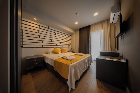 A modern hotel room with a wooden floor, a bed with a yellow bedspread, a bedside table, a dresser, and a mounted TV.