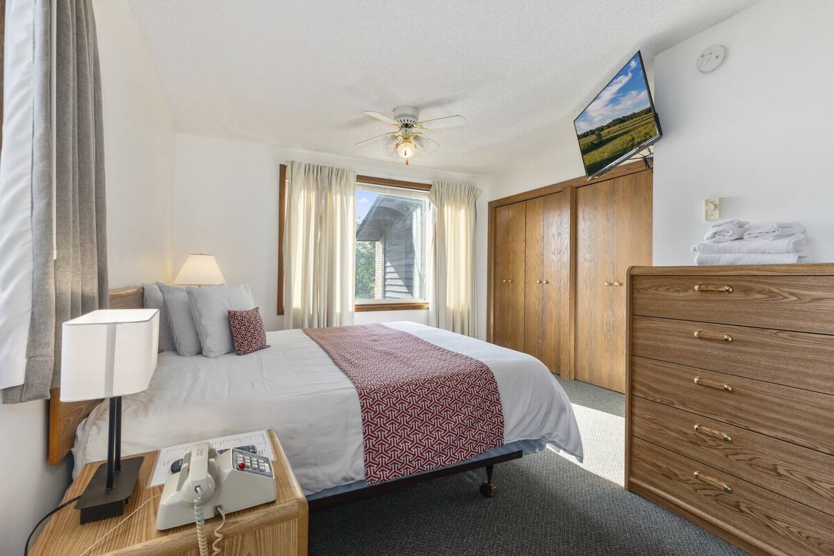 A hotel room with a bed, a desk with a telephone, a wardrobe, a television on the wall, and a window with curtains.