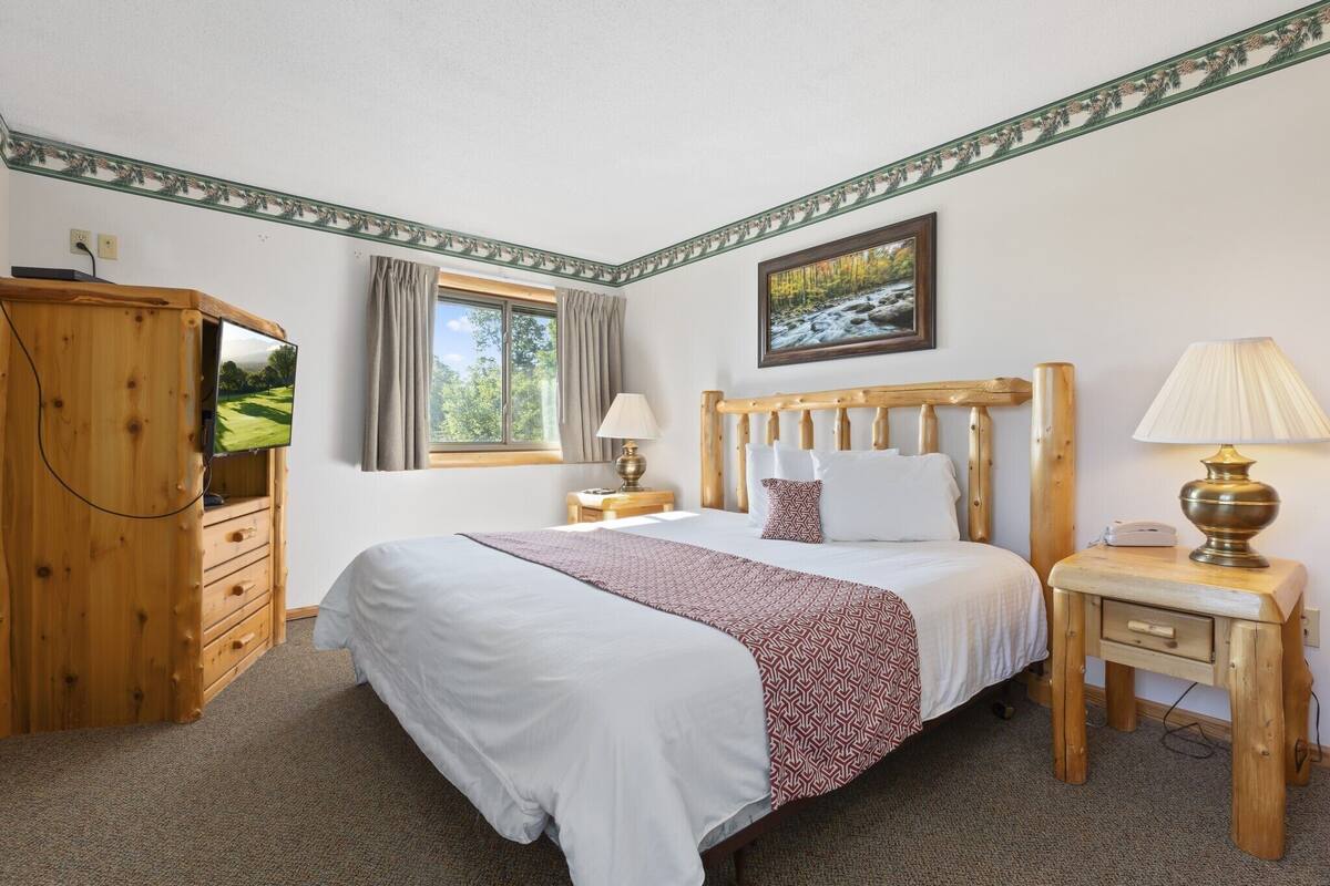 A bedroom with a large bed, wooden headboard, a nightstand with a lamp, a wooden dresser, a window with curtains, and a painting on the wall.
