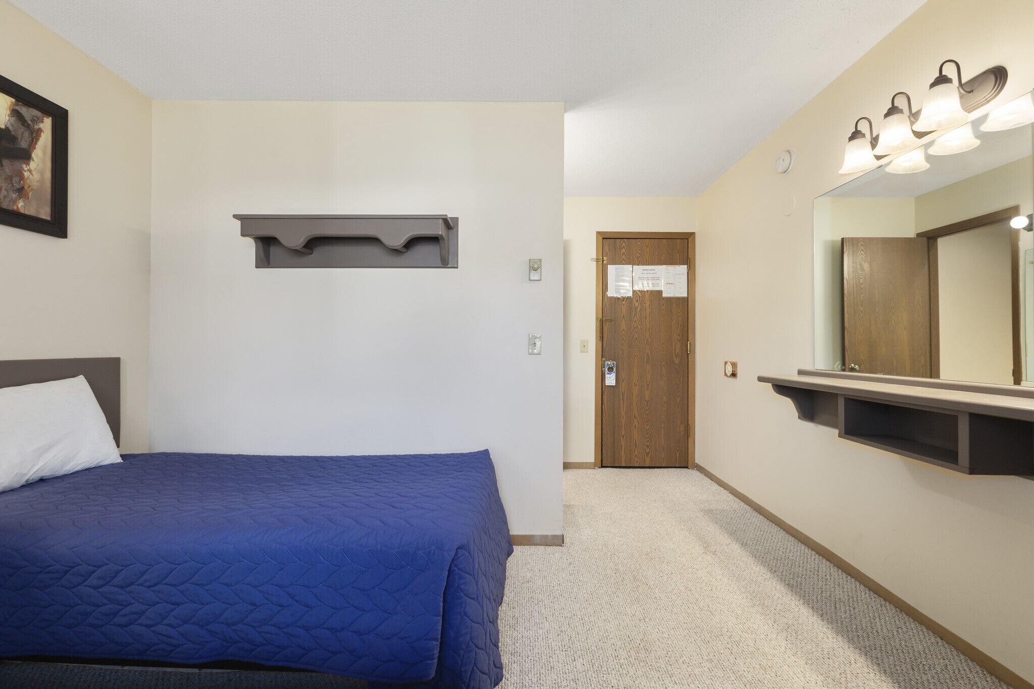 A hotel room with a bed, a wooden door, a mirror with lights, and a small shelf.
