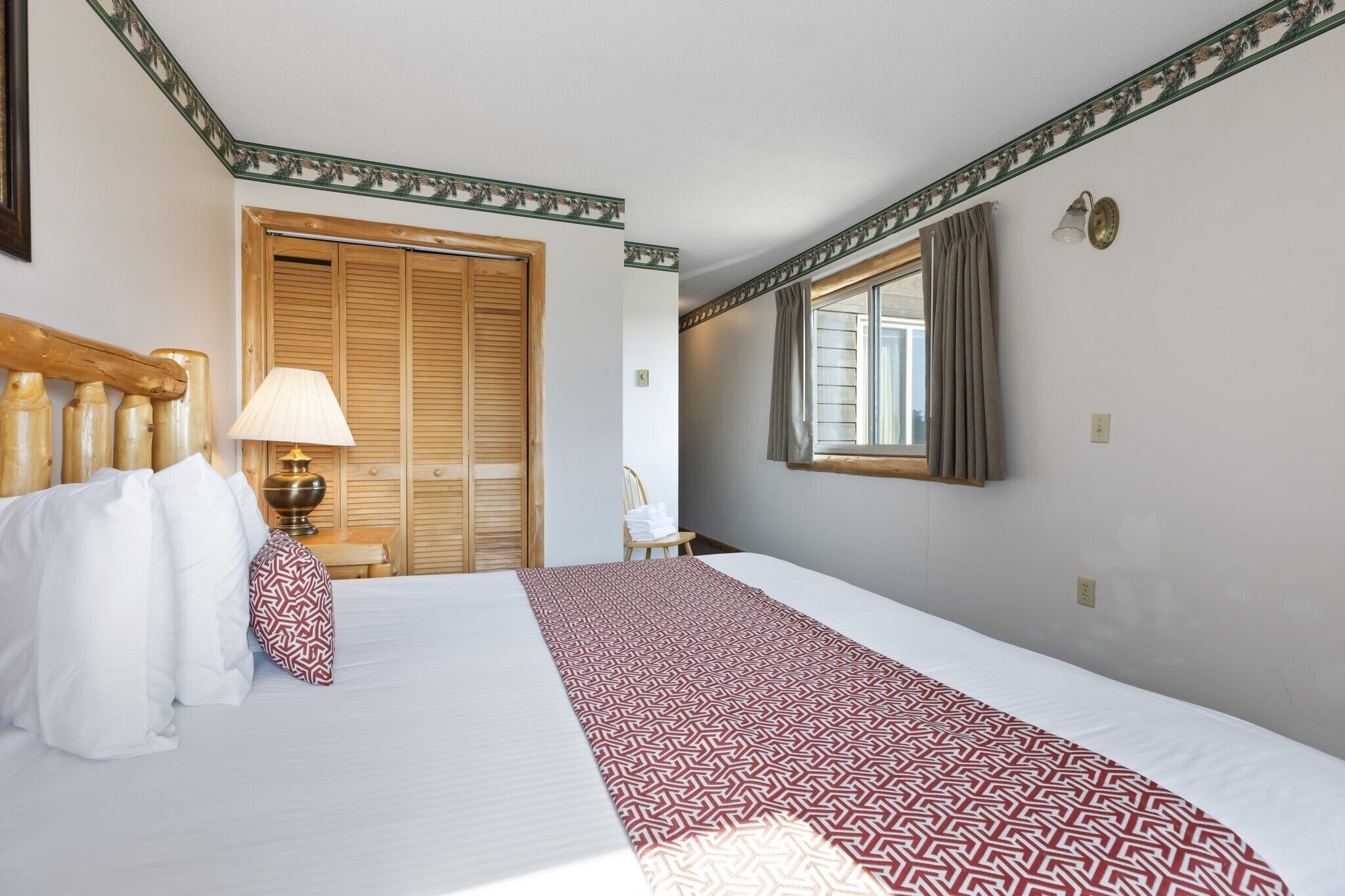 A hotel room with a large bed, a wooden headboard, a bedside lamp, a window with curtains, and a wardrobe with a mirror.