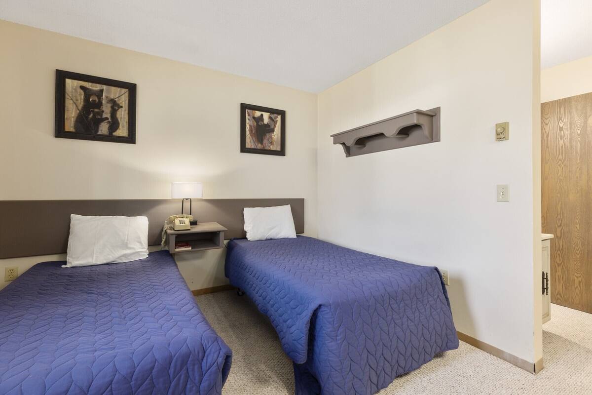 A hotel room with two beds, a bedside table with a lamp, and two framed pictures on the wall.
