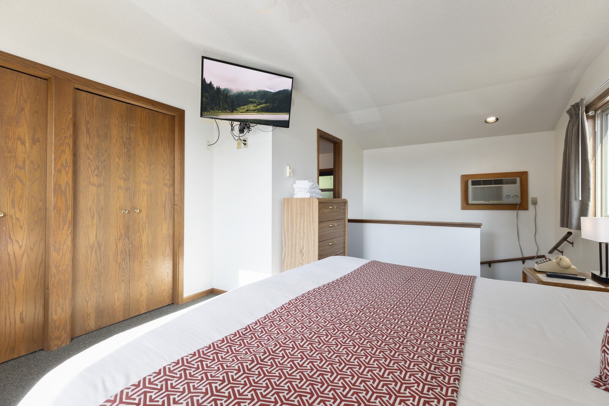 Habitación de hotel con una cama grande, armario de madera, televisor montado en la pared, escritorio con lámpara y ventana con cortinas.