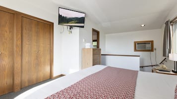 Habitación de hotel con una cama grande, armario de madera, televisor montado en la pared, escritorio con lámpara y ventana con cortinas.