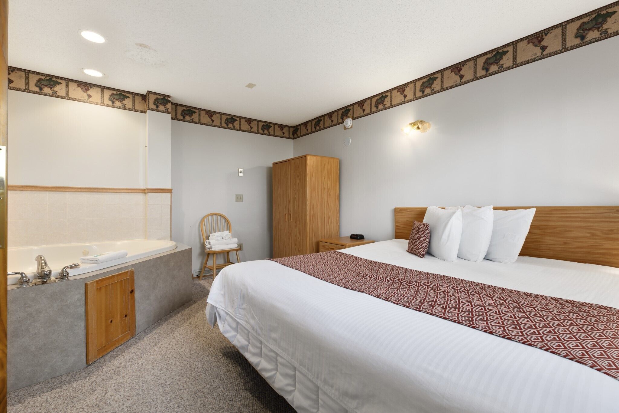 A hotel room with a large bed, a wooden wardrobe, a wooden chair, a bathroom with a bathtub, and a mirror.