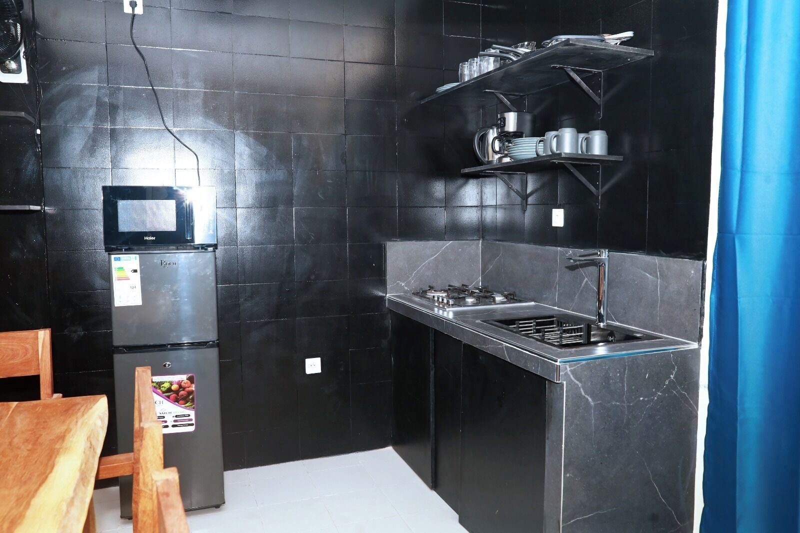 A compact kitchen with black tiled walls, a built-in microwave, a refrigerator, a gas stove, and shelves with kitchenware.