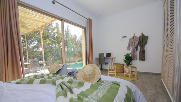 A bedroom with a large window, a bed with a striped blanket, a chair, a small table, and a plant.