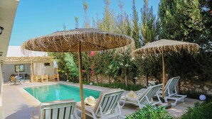 A poolside area with lounge chairs and thatched umbrellas, surrounded by greenery and a clear sky.