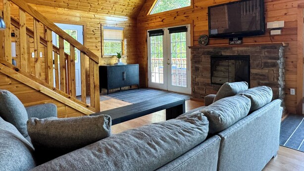A cozy cabin interior with a wooden ceiling, a stone fireplace, a television mounted above it, and a large gray sofa. - Spectacular Mountain Views. Peaceful Solitude. Rustic cabin with modern comforts. 90 mins from DC (Rileyville)