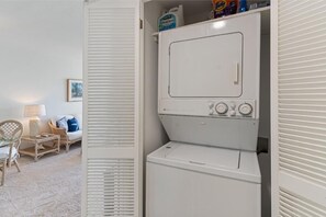 A laundry room with a front-loading washer and dryer, a wicker chair, a small table, and a framed picture on the wall. - Royal Mauian, #405 2 Bedroom Condo (Kihei)
