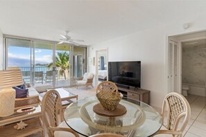 A modern living room with a glass table, wicker chairs, a flat-screen TV, and a balcony with a view of the ocean. - Royal Mauian, #405 2 Bedroom Condo (Kihei)