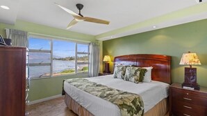 A bedroom with a bed, two bedside lamps, a ceiling fan, a window with a view of the water, and a wooden nightstand. - Royal Mauian, #207 2 Bedroom Condo (Kihei)
