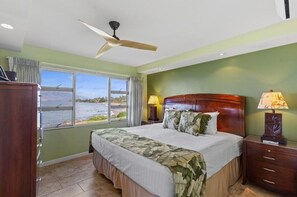 A bedroom with a bed, two bedside lamps, a ceiling fan, a window with a view of the water, and a wooden nightstand. - Royal Mauian, #207 2 Bedroom Condo (Kihei)