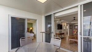 A glass-enclosed balcony with a round table and chairs, a sliding glass door leading to a bedroom, and a view into a living area with a television and a ceiling fan. - Royal Mauian, #207 2 Bedroom Condo (Kihei)