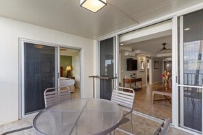A glass-enclosed balcony with a round table and chairs, a sliding glass door leading to a bedroom, and a view into a living area with a television and a ceiling fan. - Royal Mauian, #207 2 Bedroom Condo (Kihei)