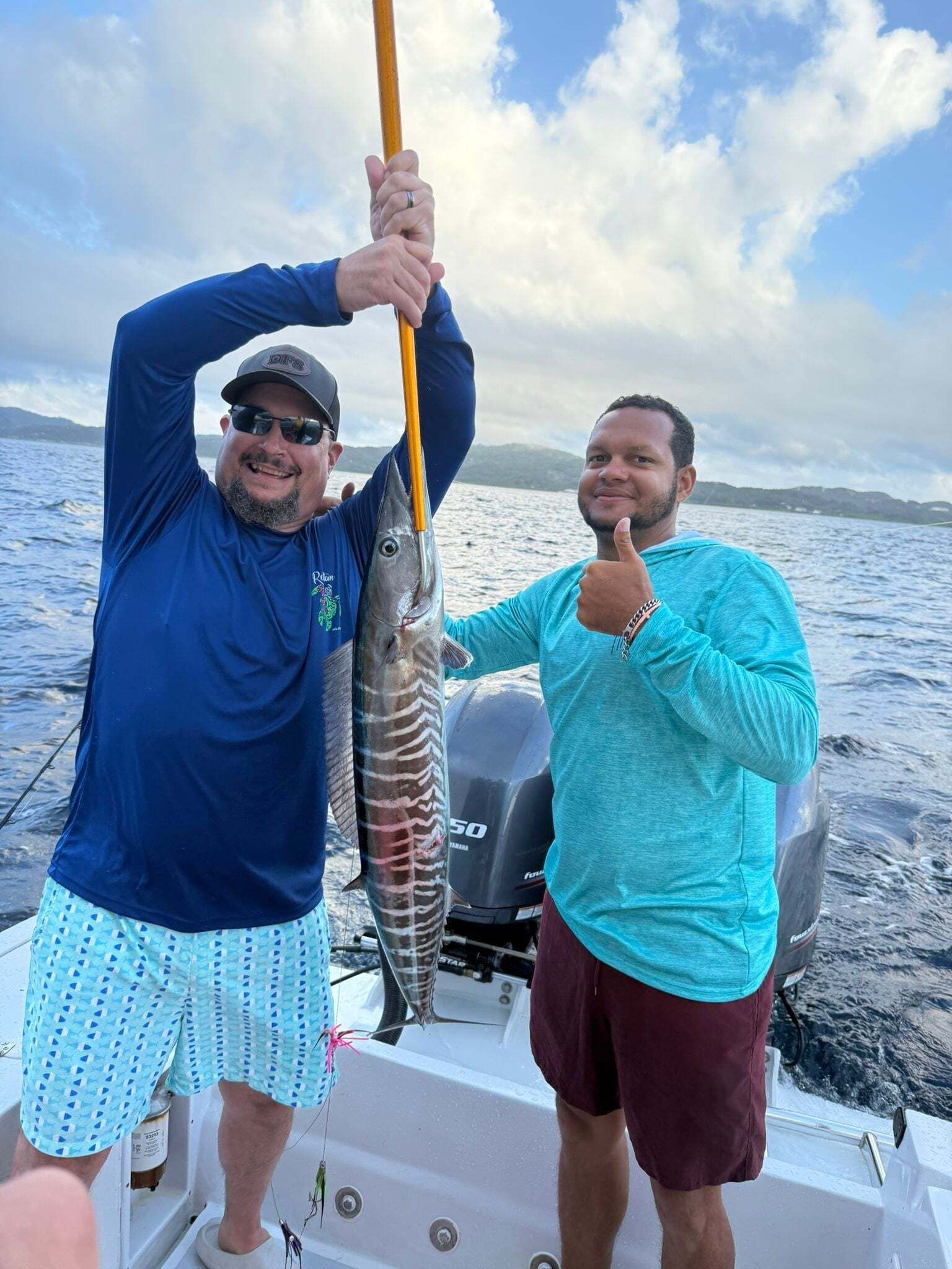 Two individuals on a boat holding a fish, with a body of water and distant land in the background.
