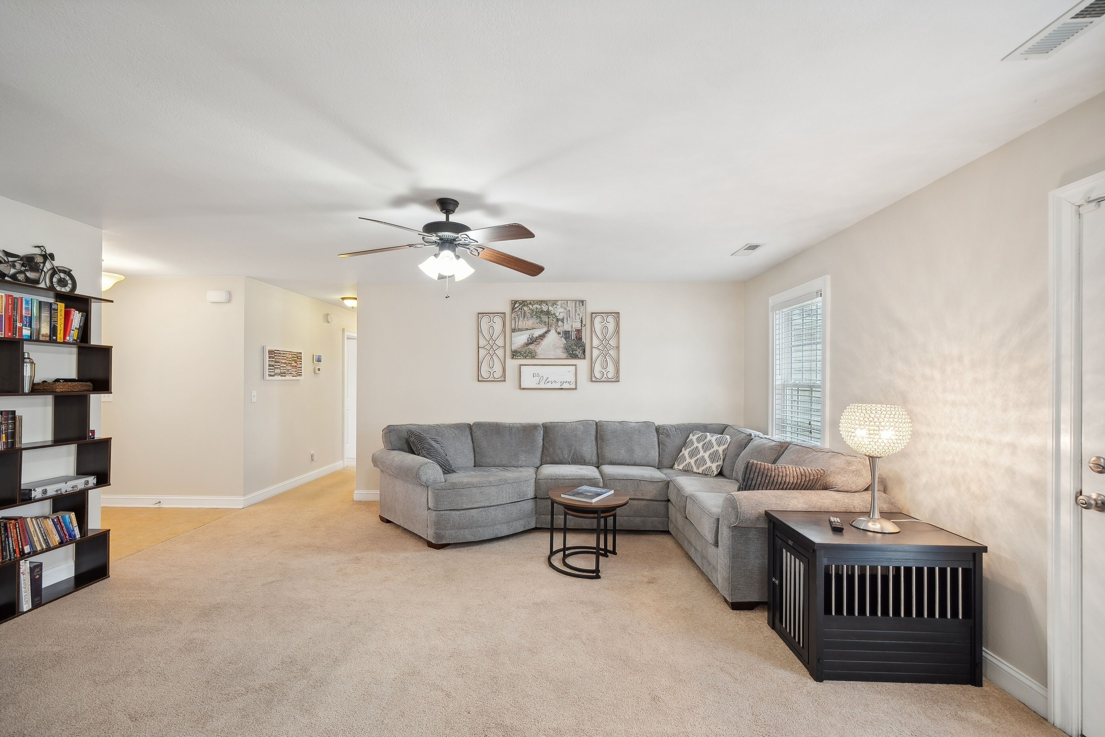 A living room with a sectional sofa, a small round table, a bookshelf, a floor lamp, and a ceiling fan.