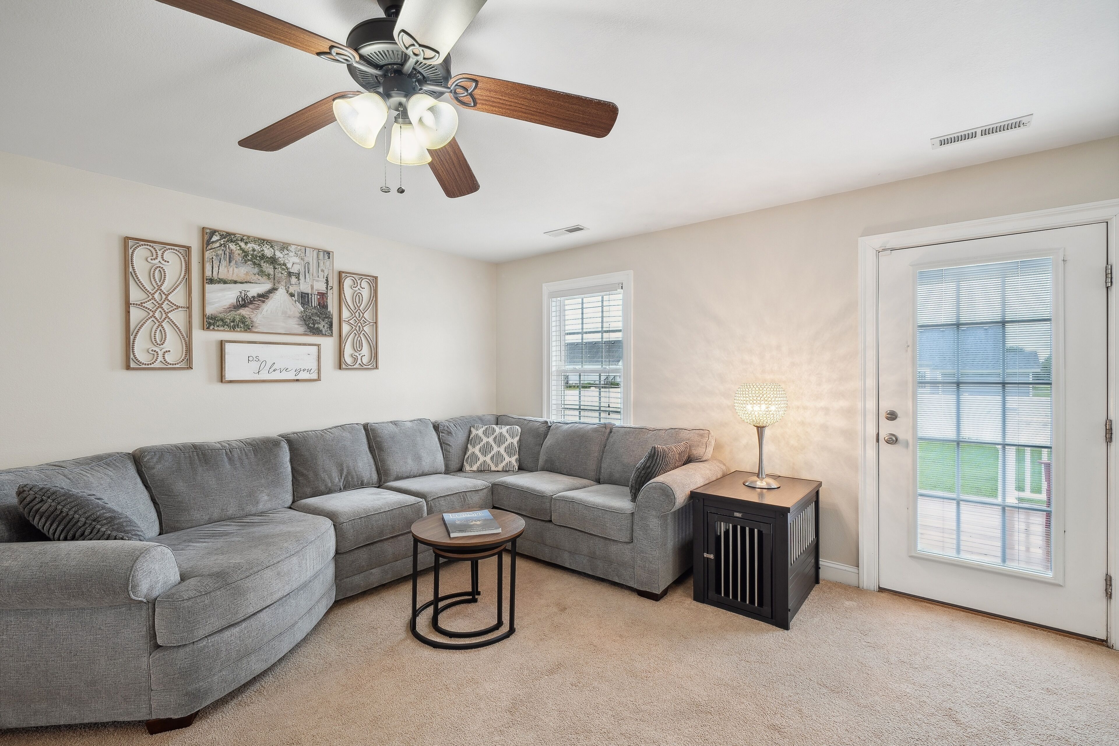 A living room with a gray sectional sofa, a small round coffee table, a ceiling fan, and a door with a view to the outside.