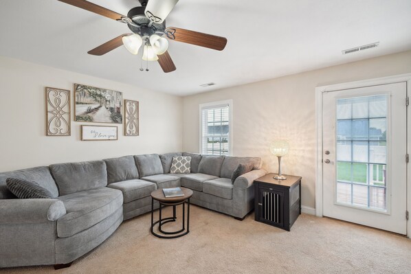 A living room with a gray sectional sofa, a small round coffee table, a ceiling fan, and a door with a view to the outside.