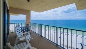 Ein Balkon mit weißen Stühlen und einem Tisch, der auf einen Strand mit Sonnenschirmen und Menschen blickt.
