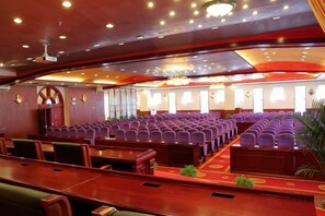 Une salle de conférence avec des rangées de chaises et de tables, une scène et un plafond doté d’un éclairage encastré.