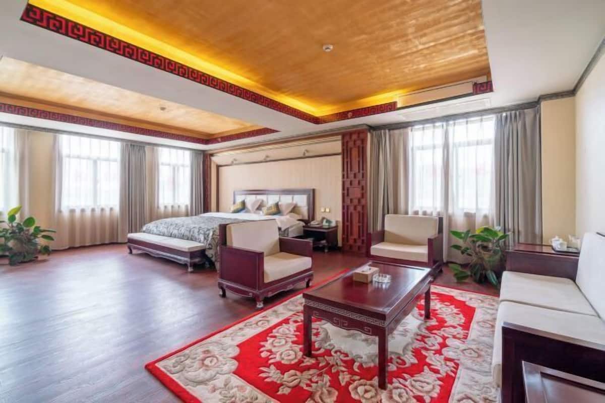 Hotelkamer met een groot bed, twee fauteuils, een salontafel, een bureau en een rood vloerkleed met bloemen.
