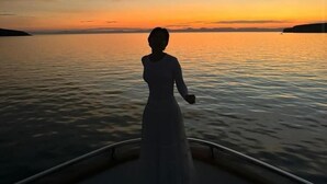 Pitkän mekon päällään pitävä henkilö seisoo veneessä ja katsoo auringonlaskua tyynen veden yllä.
