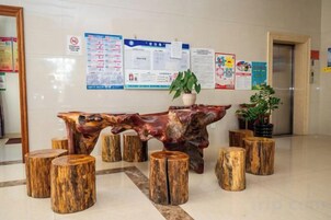Un área de recepción con un banco y taburetes de madera, una planta en maceta y carteles informativos en la pared.