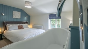 A bedroom with a large bed, a white bathtub, a window with a view of greenery, and a decorative wall piece. - Jasmine Cottage, Sedgeford, Norfolk (Sedgeford)
