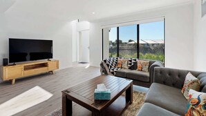 A modern living room with a flat-screen TV, wooden entertainment unit, a coffee table, a grey sofa, and a large window with a view of a residential area. - 4-13 Central Dr Victor Harbor - Wi-Fi and Linen (Victor Harbor)