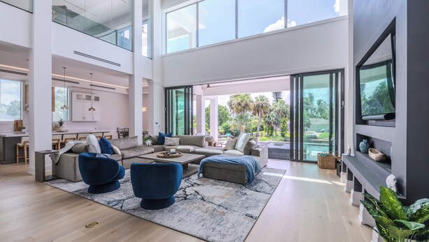 A spacious living area with a large sofa, blue armchairs, and a coffee table. There is a kitchen area with a dining table and chairs visible in the background. - Free Golf Cart Aug & Sep! Waterfront Mansion (Naples)
