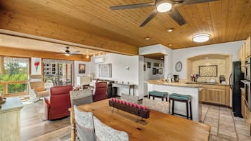 A spacious living area with a wooden ceiling, a dining table with chairs, a kitchen area, and a view through large windows.