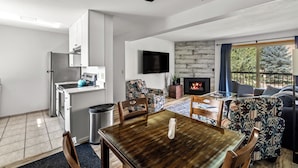 Un salon moderne avec une cheminée, un coin repas avec une table en bois et une cuisine équipée d’un réfrigérateur et d’un téléviseur.