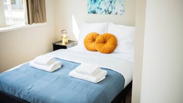 A neatly made bed with blue bedding, two folded white towels, and an orange pillow.