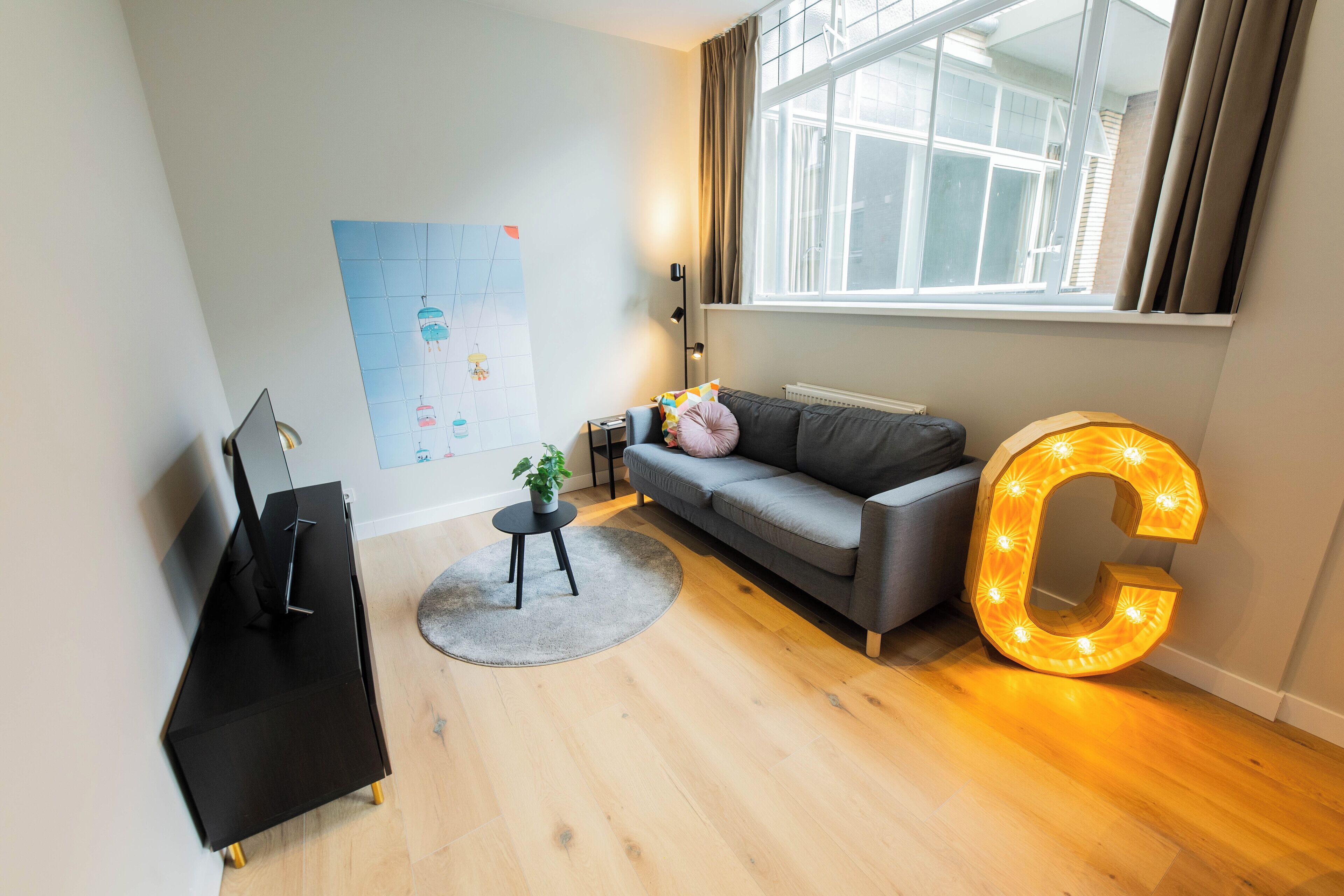 A modern living room with a grey sofa, a black TV stand, a round rug, a large illuminated 'C' decoration, and a window with curtains.