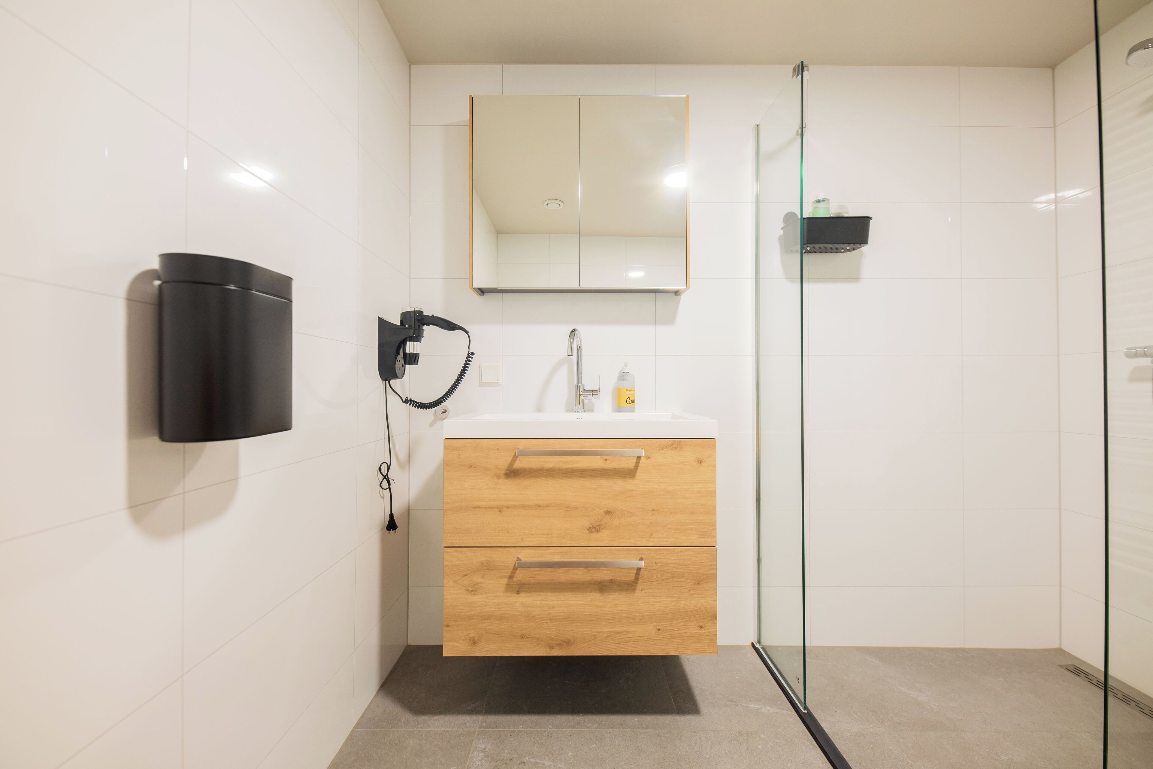 Ein modernes Badezimmer mit weiß gefliesten Wänden, einem hölzernen Waschtisch mit zwei Schubladen, einem Spiegel über dem Waschtisch, einem schwarzen Seifenspender und einer Duschkabine aus Glas.