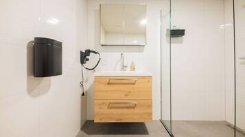A modern bathroom with a white tiled wall, a wooden vanity with two drawers, a mirror above the vanity, a black soap dispenser, and a glass shower enclosure.
