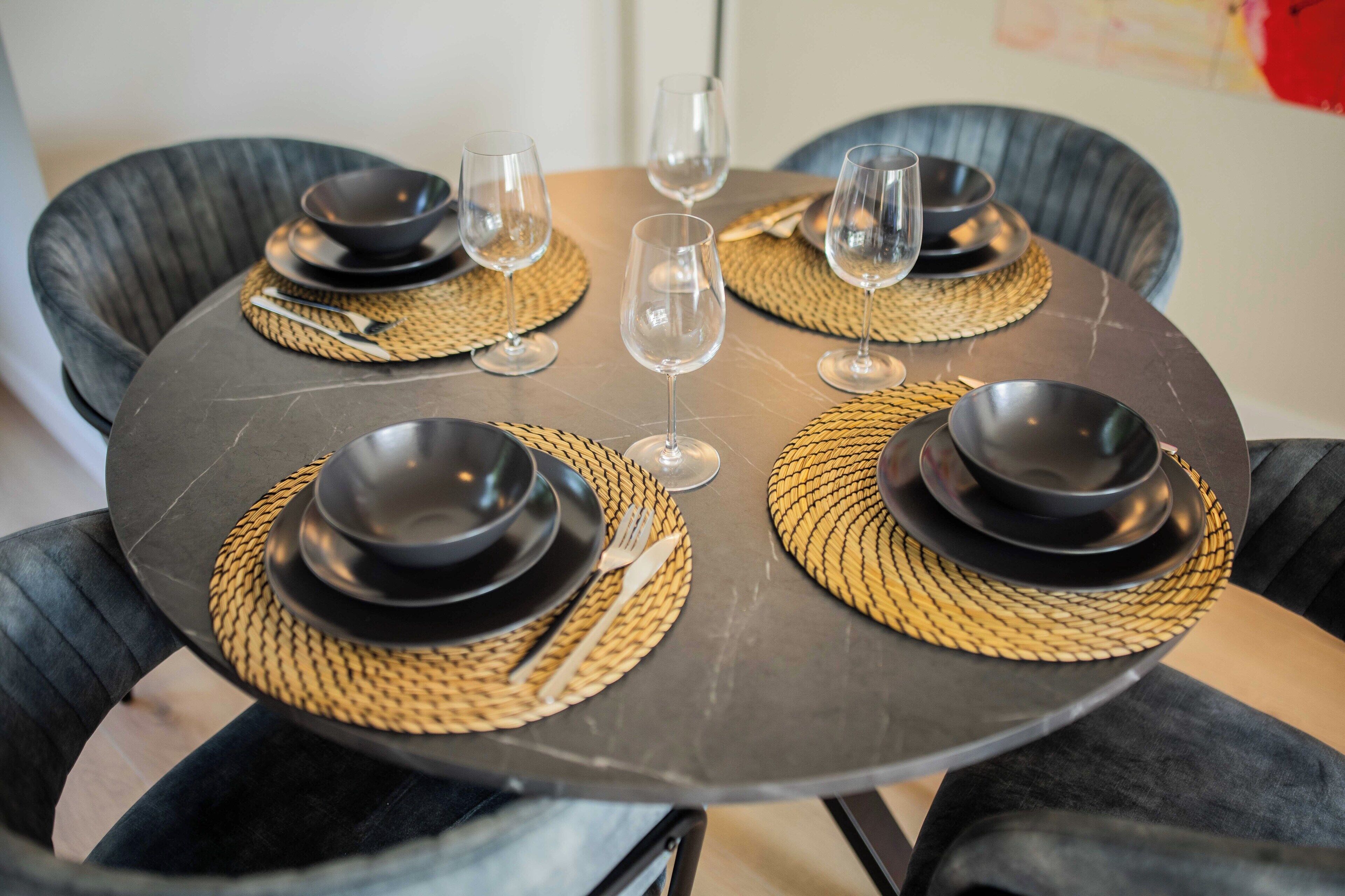 Une table de salle à manger ronde, avec des assiettes noires, des verres transparents et des sets de table en fibres tressées.