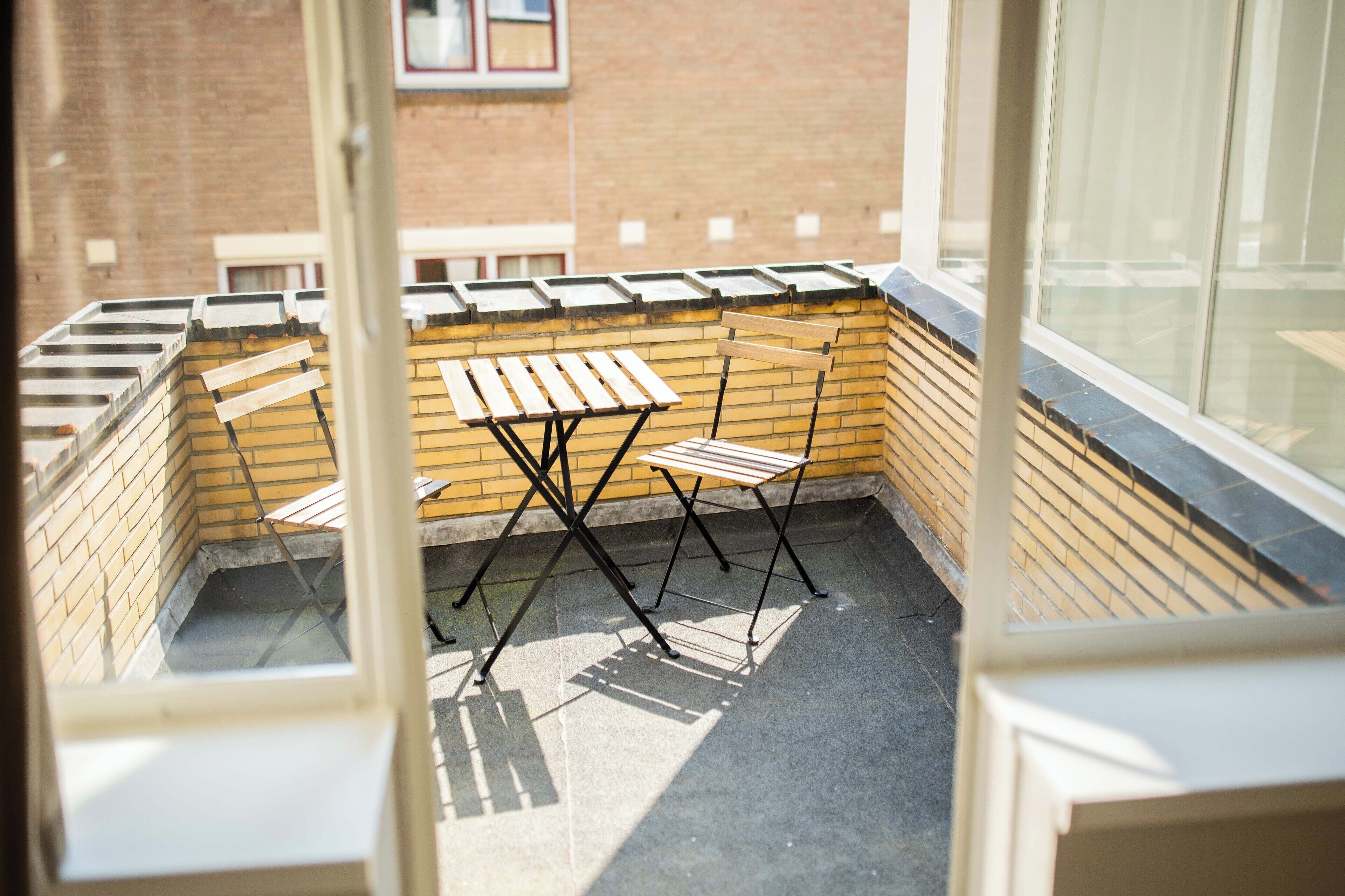A view from a window overlooking a small outdoor seating area with a table and chairs, a brick wall, and a window with blinds.