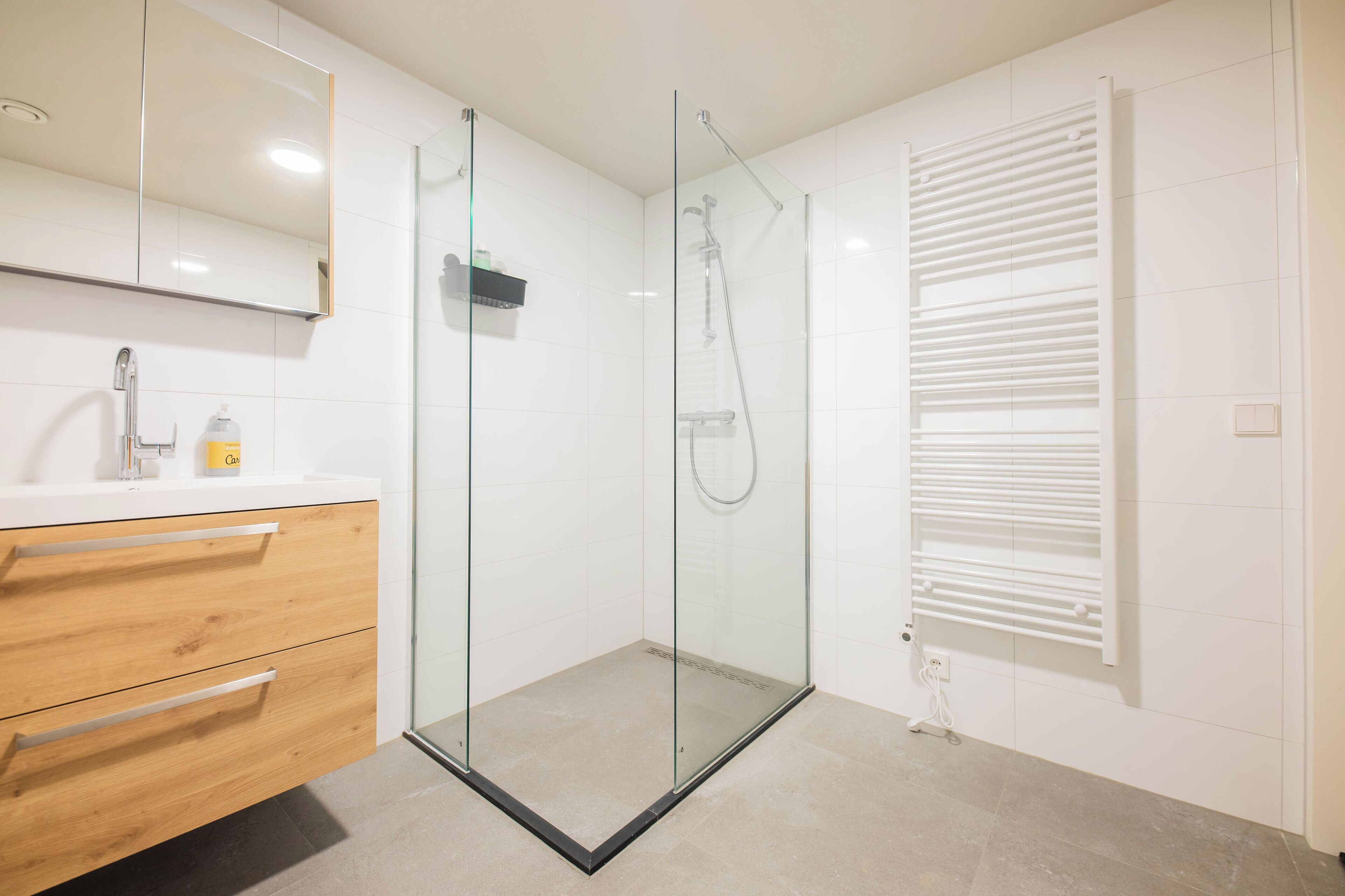 Ein modernes Badezimmer mit einer in Glas eingefassten Dusche, einem hölzernen Waschtisch mit zwei Schubladen, einem Spiegel und einer weiß gefliesten Wand.