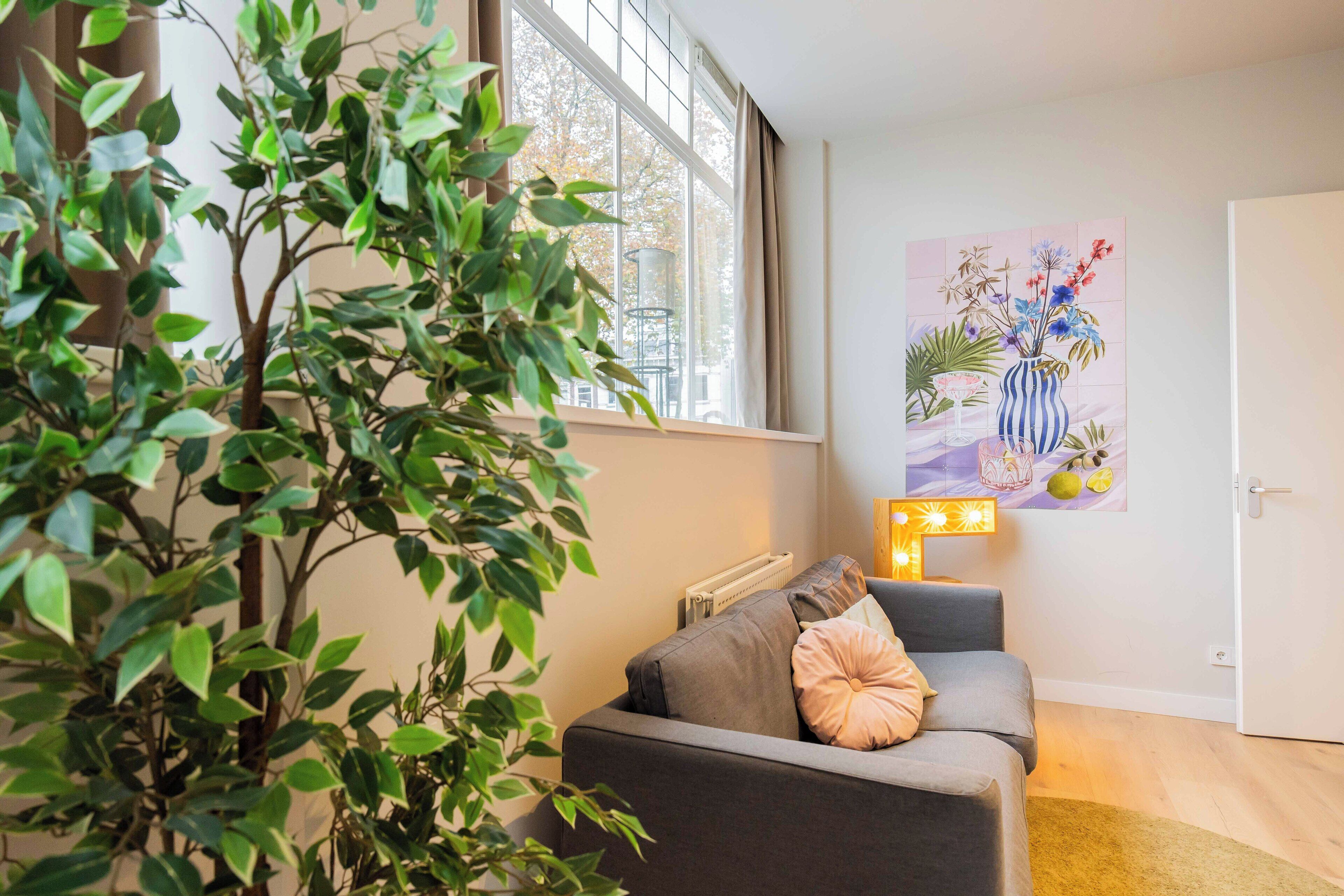 A living room with a grey sofa, a large potted plant, a window with curtains, and a wall-mounted artwork.
