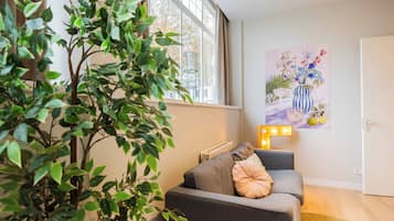 A living room with a grey sofa, a large potted plant, a window with curtains, and a wall-mounted artwork.