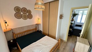 A bedroom with a bed, bedside lamps, a hanging light fixture, a wardrobe, and a view of the adjacent room.