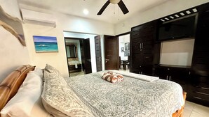 Une chambre à coucher avec un lit, un ventilateur de plafond, une télévision et une salle de bain visible à travers une porte ouverte.