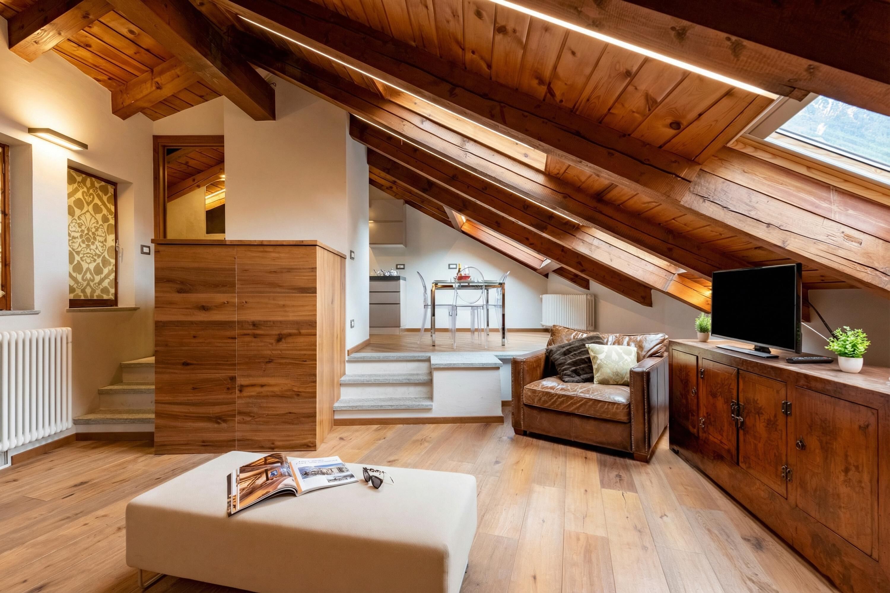 A loft-style room with wooden beams, a white sofa, a TV, and a staircase leading to an upper level.