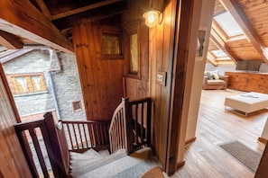 Una scala in legno conduce a una camera da letto mansardata con una finestra a lucernario, una parete in pietra e un moderno divano bianco.